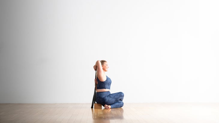 Woman with blonde hair sits on a cork block and uses a strap to practice Cow-face pose. She is wearing an ocean blue top and yoga tights.