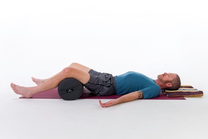 A man lies in Corpse Pose (Savasana) with a bolster under his knees