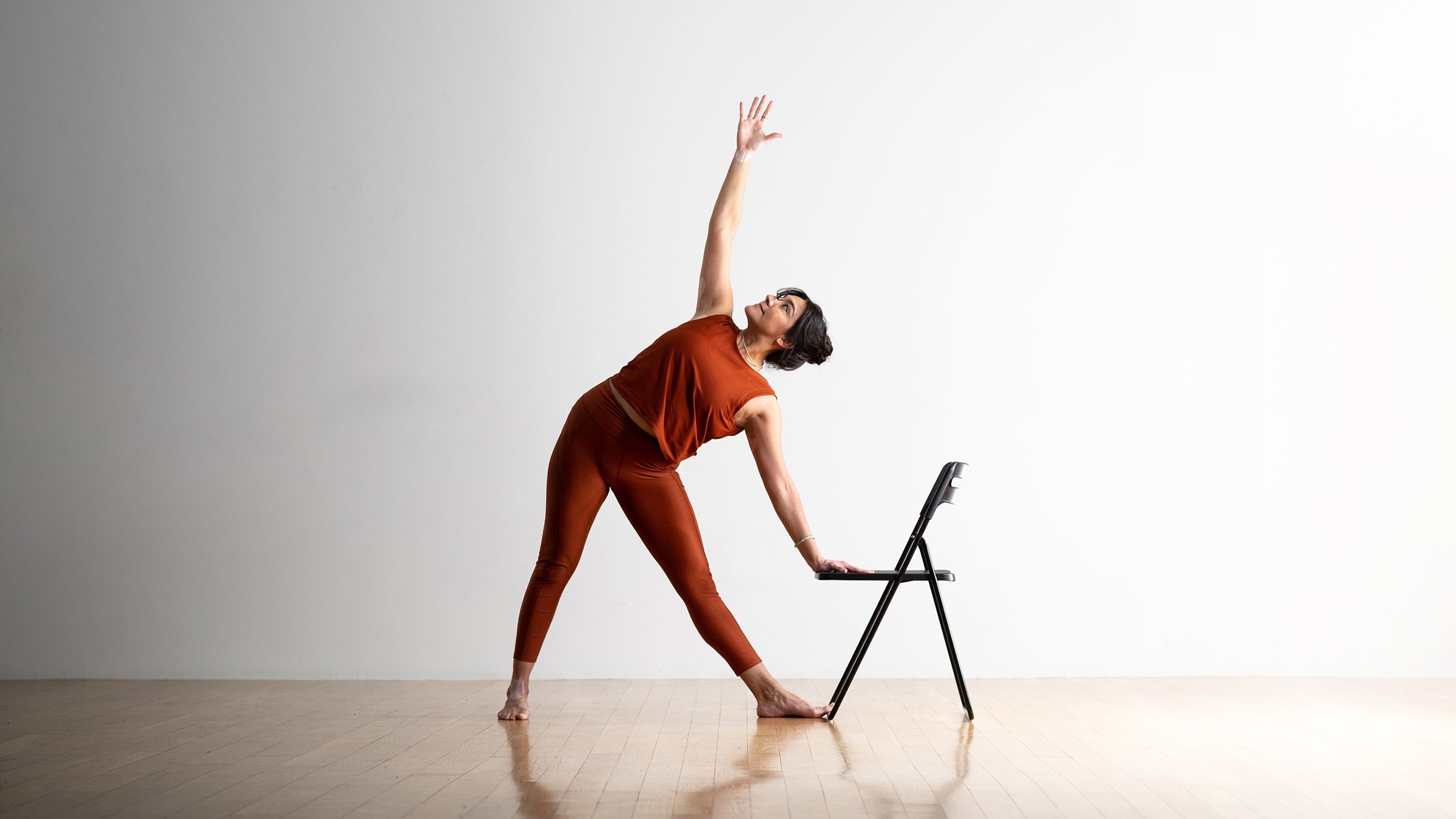 A woman in rust colored yoga clothing practices Trikonasana (Triangle Pose) with one hand on a chair. A white wall is in the background.