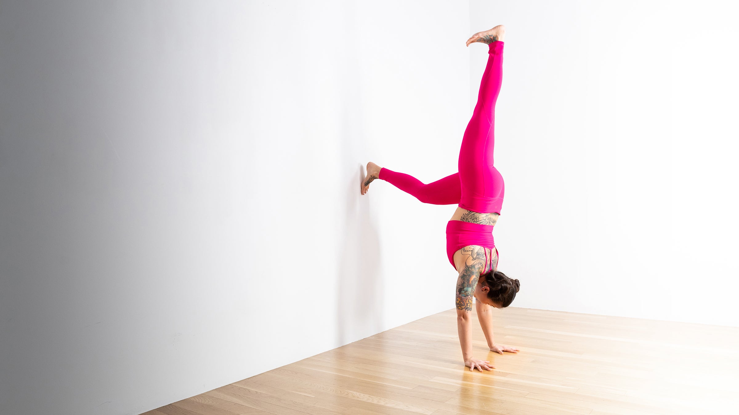 A woman in bright pink yoga clothes practices a handstand modification against a white wall.