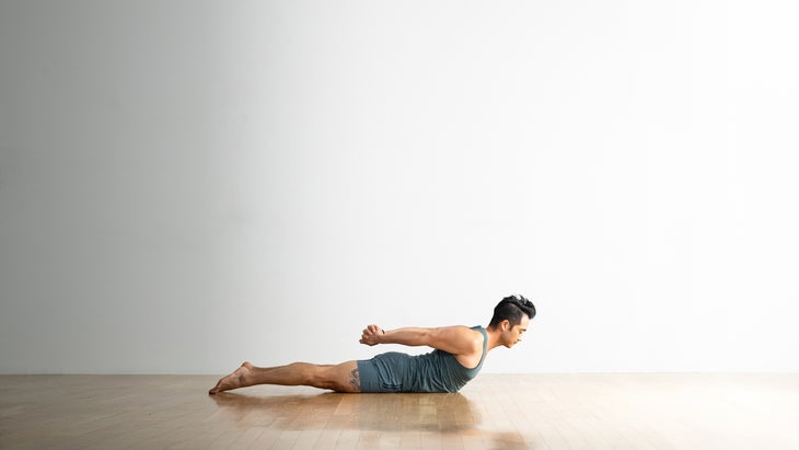 Hiro Landazuri practicing Half Locust pose with his legs on the floor, shoulders up and hands clasped behind his back
