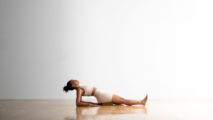 Woman practices Supta Ardha Virasana (Half Reclining Hero Pose. She draw just her right leg back into Virasana, and the other leg is extended. She leans back and props herself up on her elbows and forearms. She is a Black woman with a ponytail, wearing off whit yoga shorts and a matching top. The floor is wood and the wall behind her is white.