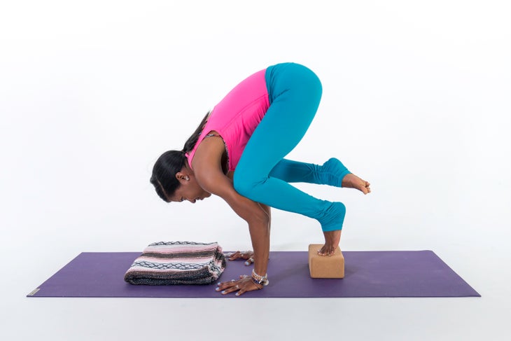 A person demonstrates a variation of Crow Pose or Crane Pose, with her foot resting on a block
