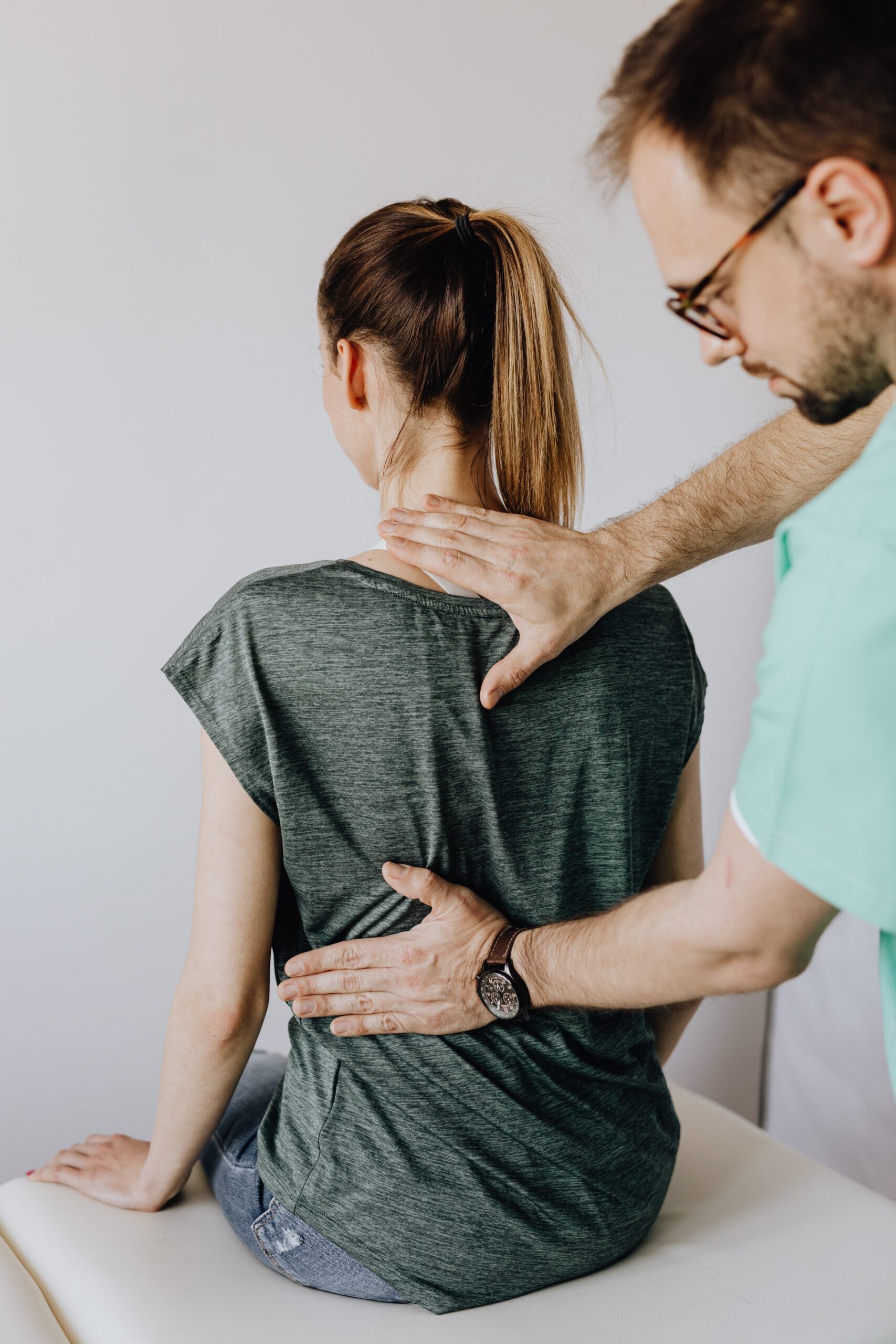 A doctor in light blue scrubs examines a the back of a woman wearing a green t-shirt.