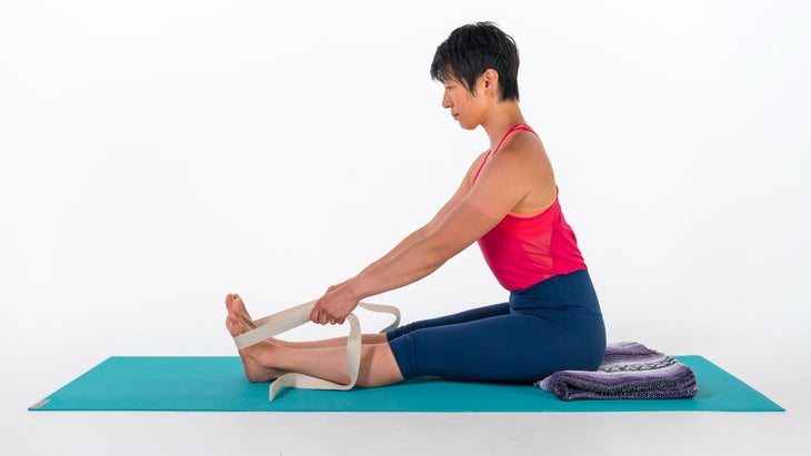 Woman demonstrates a variation of Seated Forward Bend with a blanket under her bottom and a strap around her feet
