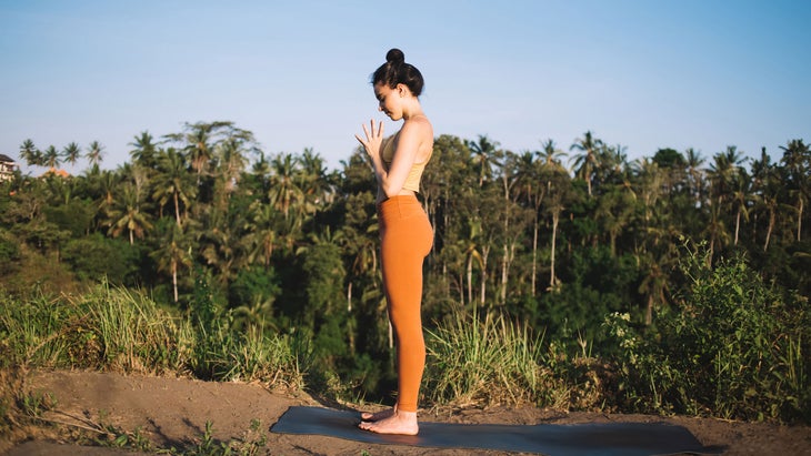 A woman stands in Tadasana with her hands at her chest while outside on her yoga mat