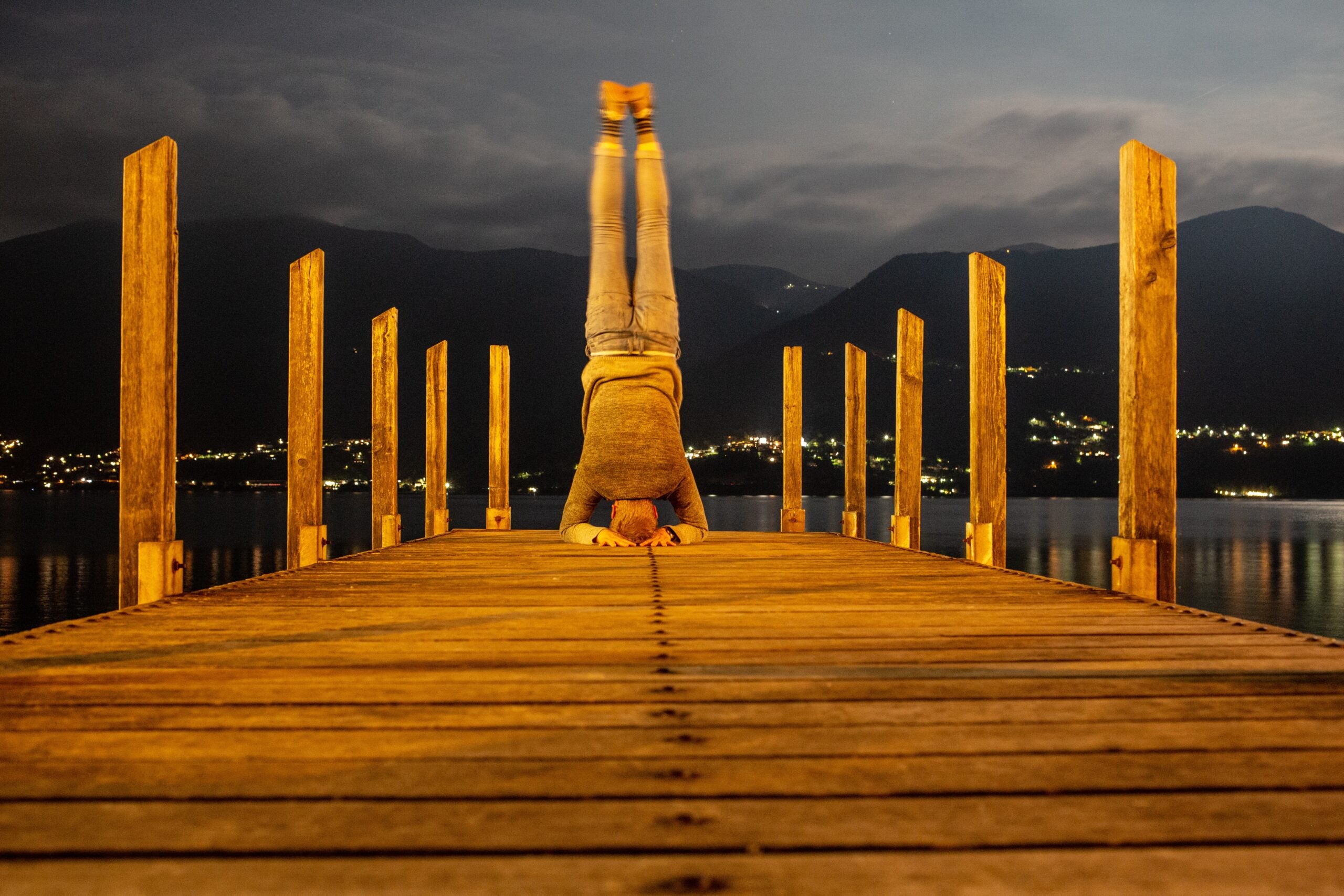 A man does a Supported Headstand at the end of a peer. It is dark dust and the pier is light brown wood. Wooden pilons line the boardwalk
