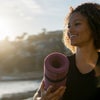 A young woman carries a yoga mat to the beach while looking off in the distance and smiling