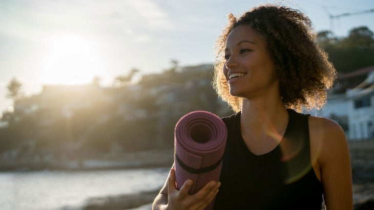 A young woman carries a yoga mat to the beach while looking off in the distance and smiling