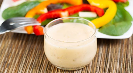 Horizontal photo of a small glass bowl filled with creamy Italian dressing with salad on white plate in background