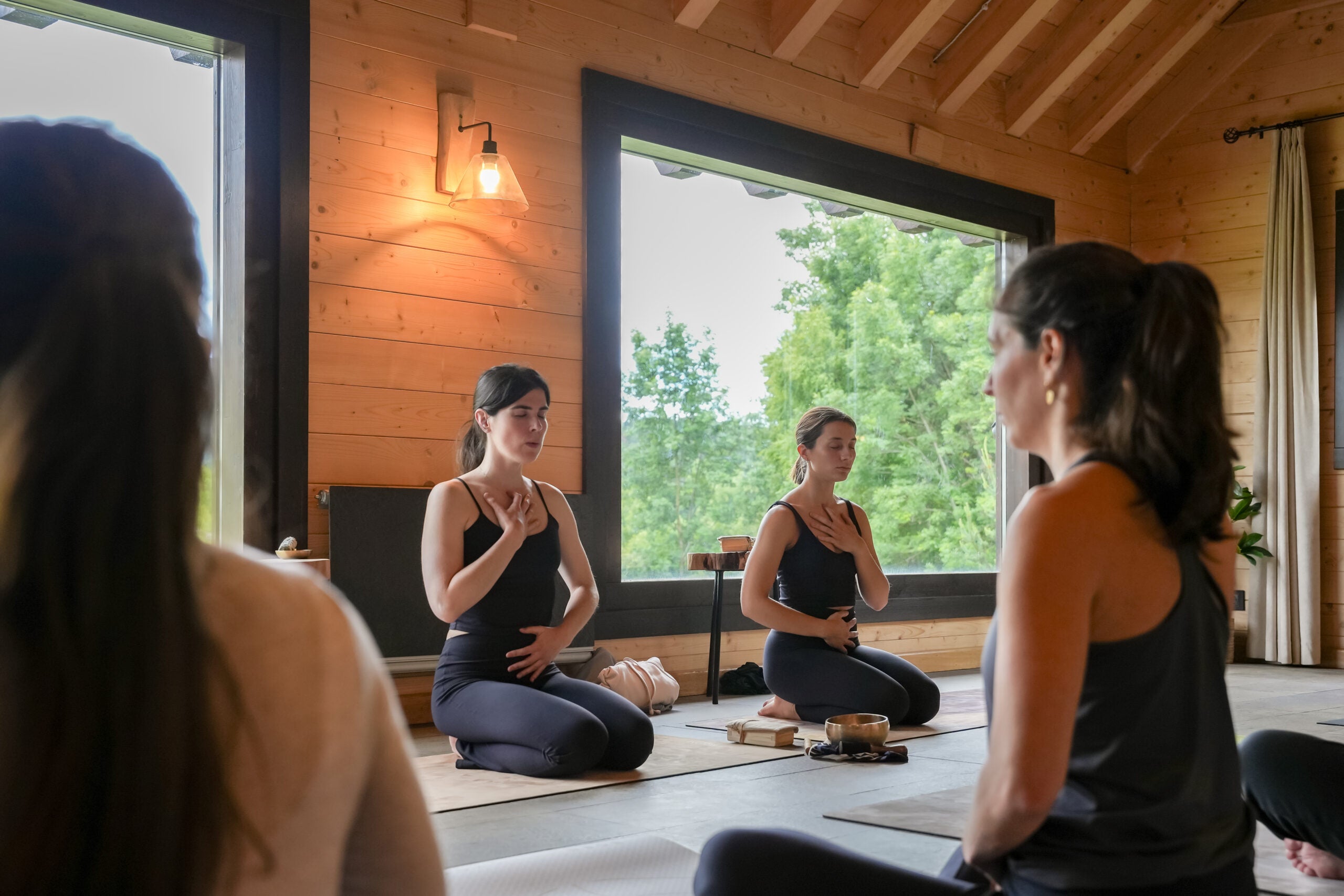 Four women in a yoga class, a window exposing green tree leaves in the background.