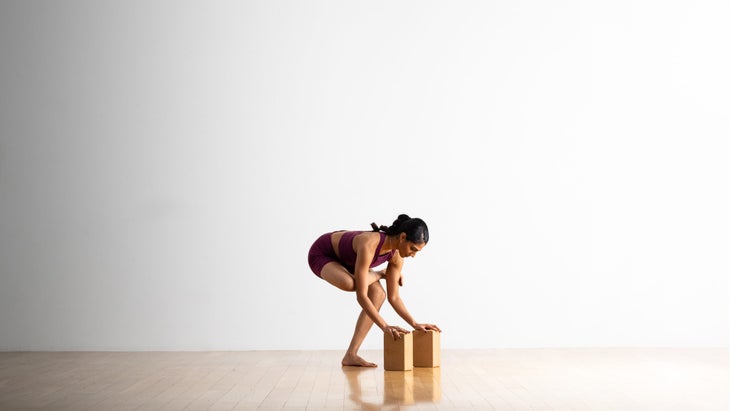 Neeti Narula practices a variation of Flying Pigeon. From a standing figure-four pose, she squats down and places her hands on blocks.
