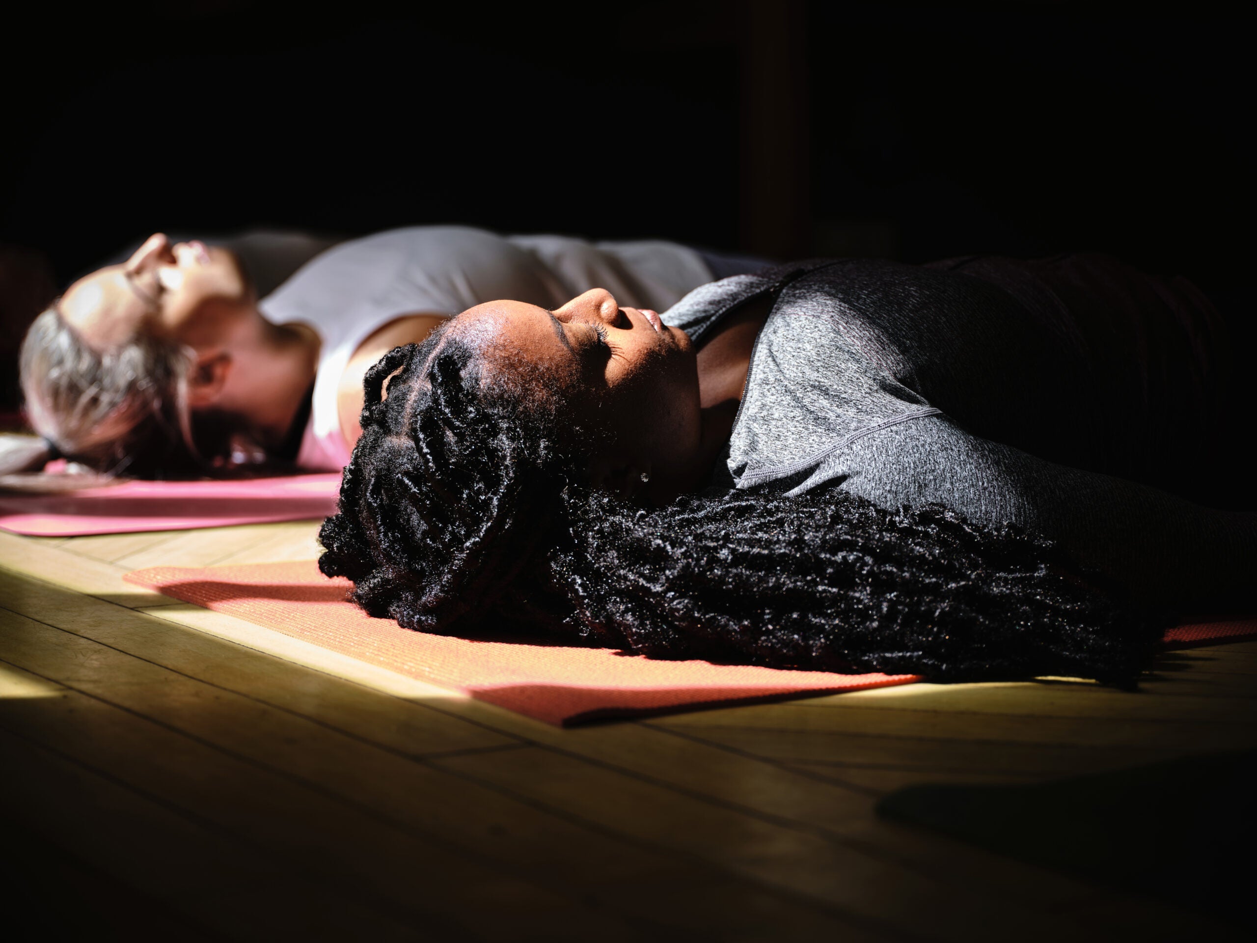 Two women laying on yoga mats in a dark room with their eyes closed.