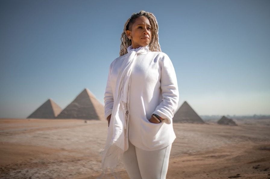 Ajax Jackson wearing a white jacket, scarf and pants, standing in front of the Pyramids in Egypt