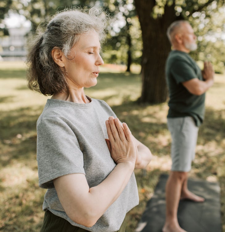 A woman with gray hair pulled back, wearing a gray t-shirt holds her hands in anjali mudra at her heart while she breaths. She is outside in a grassy area surrounded by trees. Nearby, a man with a white beard and a green t-shirt stands on a black yoga mat. His hands are in the same prayer position.
