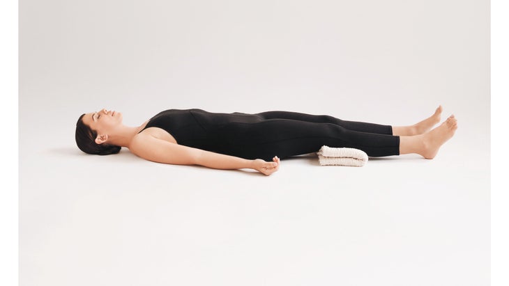 A woman wearing black demonstrates Savasana (Corpse Pose) in yoga