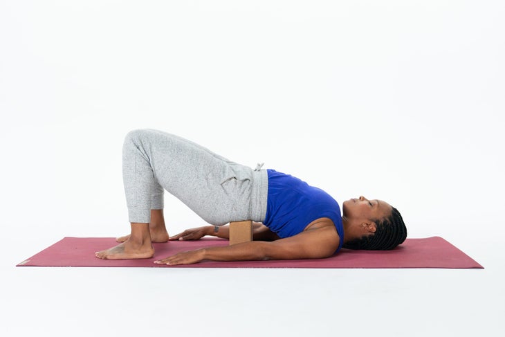 Black woman with braids, bright blue tank, gray sweatpants, Setu Bandha Sarvangasana on a block