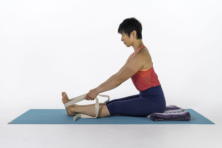 Asian woman, brown hair, red tank, blue tights, using a strap in Paschimottanasana