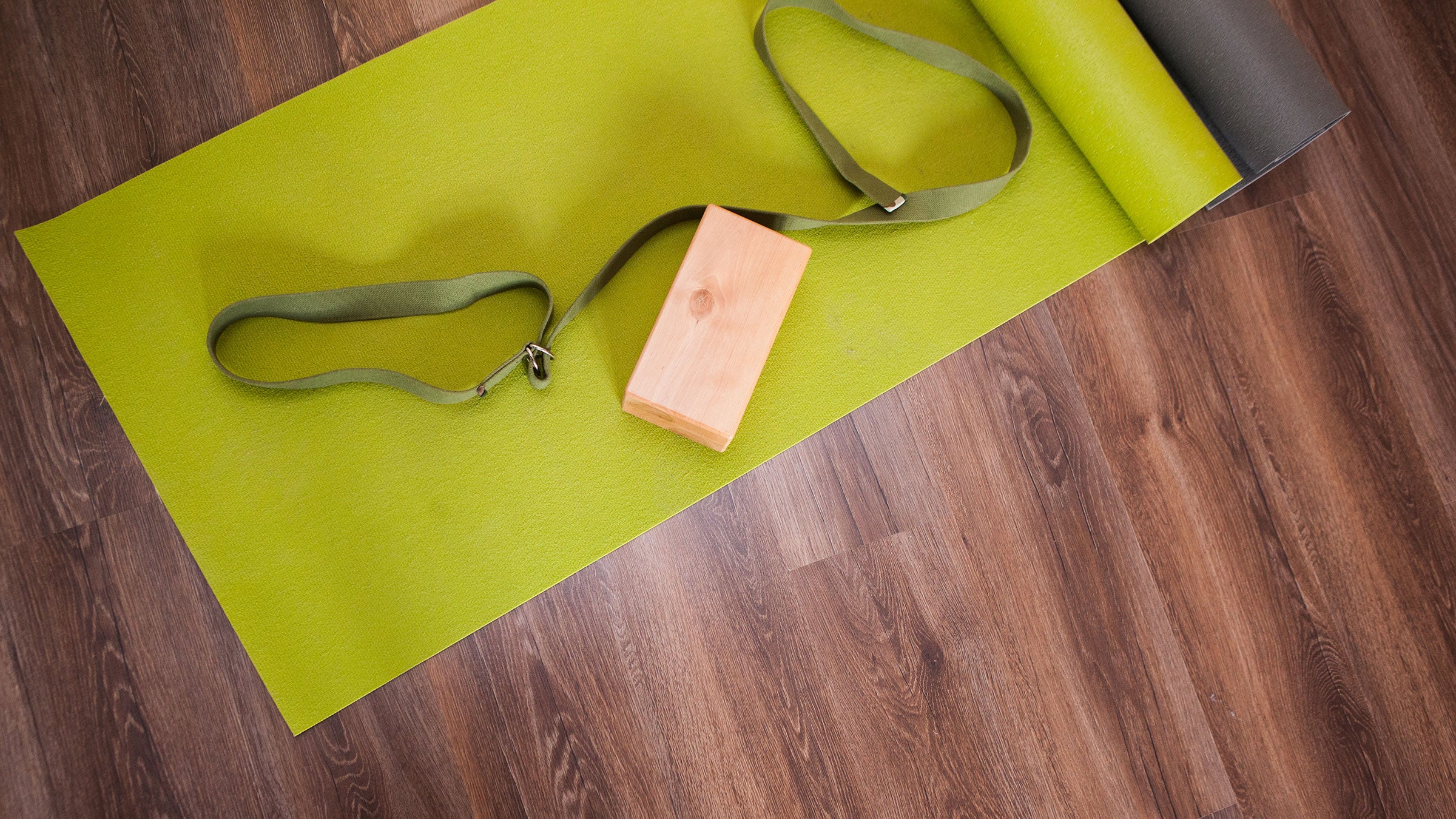 A green yoga mat, a cork block, and a stretching strap on a wood floor