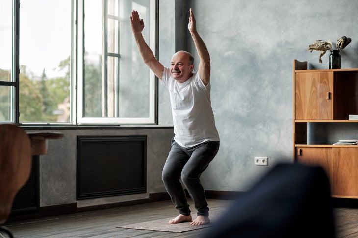 Bald, senior man in white t-shirt and gray sweat pants does Utkatasana, chair pose