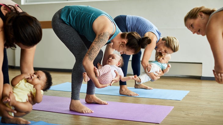 A group of women do postpartum yoga with their babies, guided by an instructor