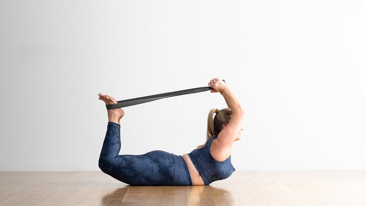 A person demonstrates a variation of Dhanurasana (Bow Pose) in yoga, using a strap