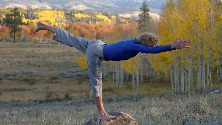A woman practices Warrior III in front of the fall aspen trees