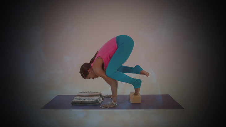 Woman wearing bright pink shirt and teal tights does Crown Pose with a cork block under her feet