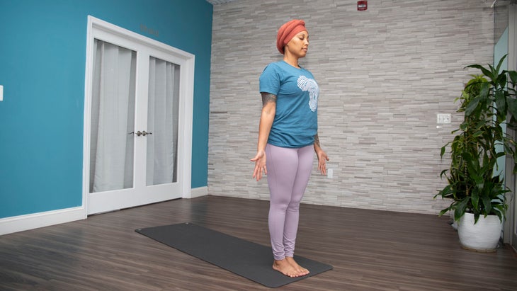 Dana Smith Stands in Tadasana wearing a teal shirt and lavender yoga pants. The Wall behind her is teal with a white framed glass door. the other wall is gray brick and there is a tall plant on the right of the photo