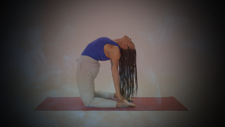 A woman with long braids, wearing a royal blue shirt and gray pants practices Camel Pose