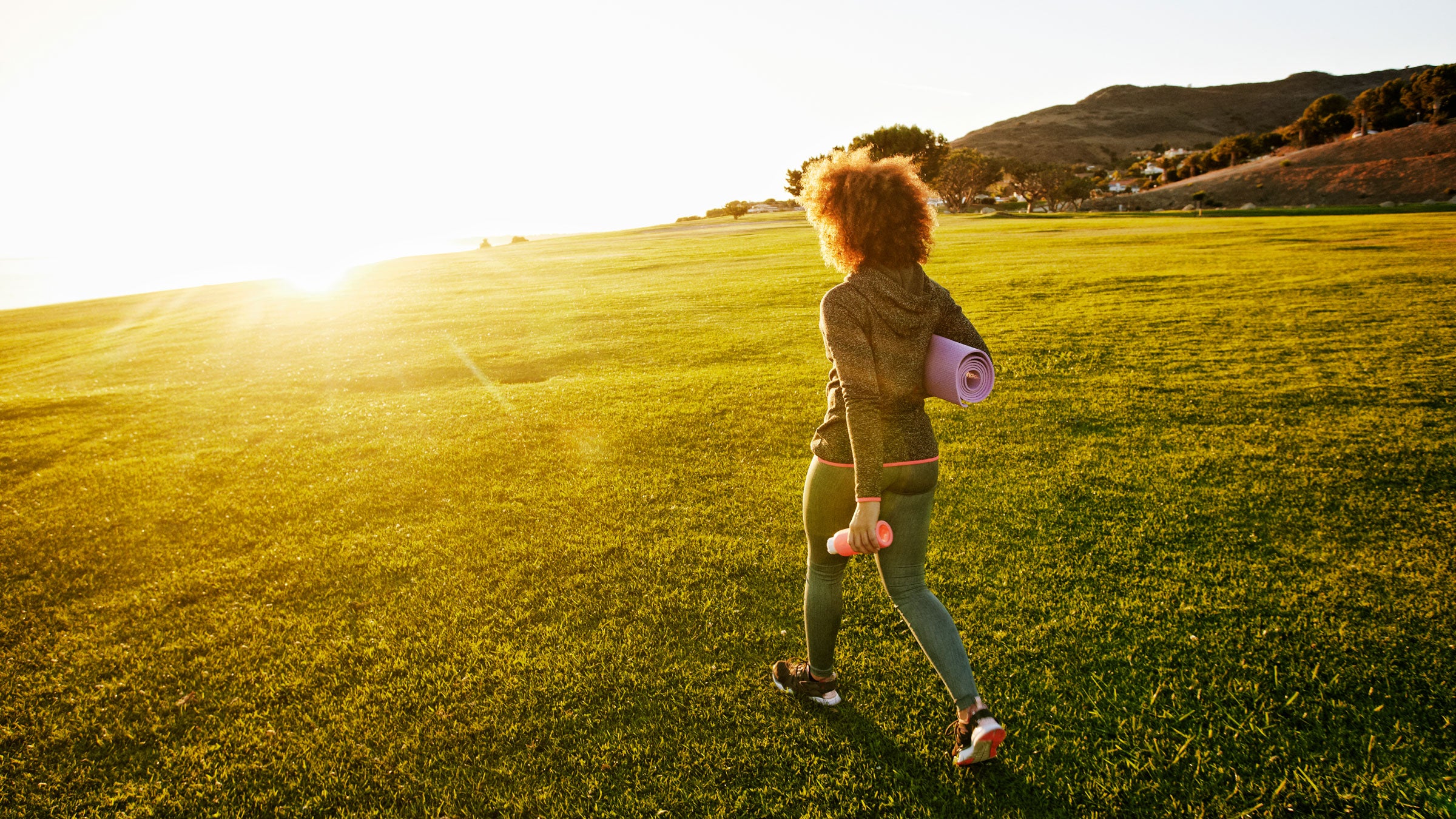 Woman walks across a grassy field with a yoga mat