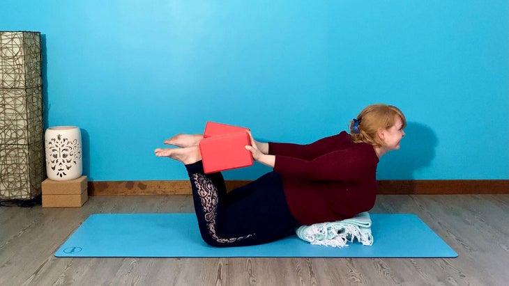A person demonstrates supported Dhanurasana (Bow Pose) using blocks to press into the ankles