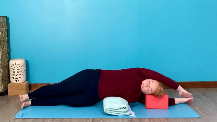 A person demonstrates supported side-lying pose with a yoga blanket and block
