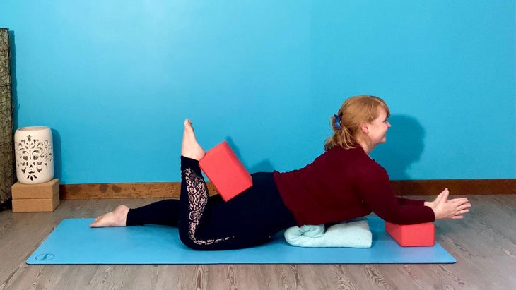 A woman demonstrates a supported Sphinx Pose with a hamstring curl using a block