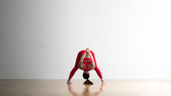 Soozie Kinstler practices a variation of a Wide-Legged Standing Forward Fold. She clasps her hands behind her back and lifts them away from her body and toward the ceiling.