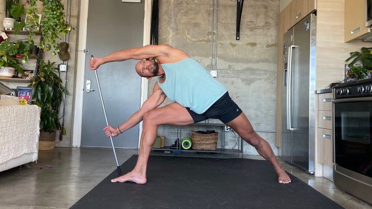 Rocky Heron in Parsvakonasana with a dowel . Yoga teacher Rocky Heron wears a light blue tank and dark shorts. He is standing on a dark/black mat with a concrete wall in the background.