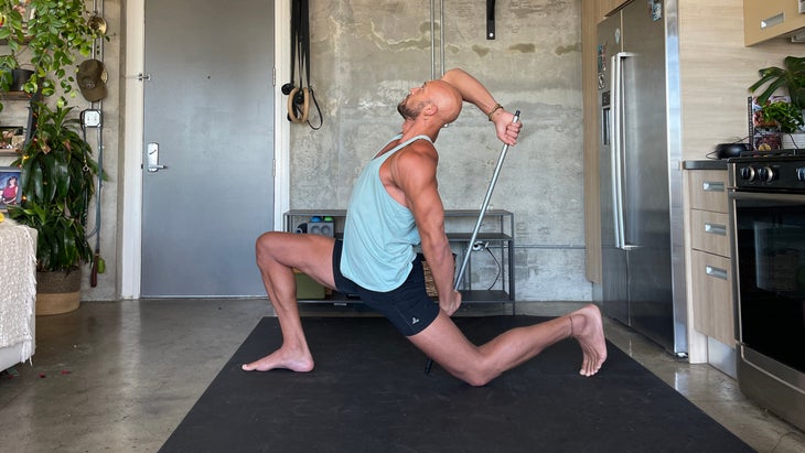 Anjaneyasana with a dowel. Yoga teacher Rocky Heron wears a light blue tank and dark shorts. He is standing on a dark/black mat with a concrete wall in the background.