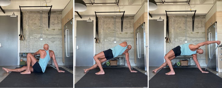 Wild Thing. Yoga teacher Rocky Heron wears a light blue tank and dark shorts. He is standing on a dark/black mat with a concrete wall in the background.