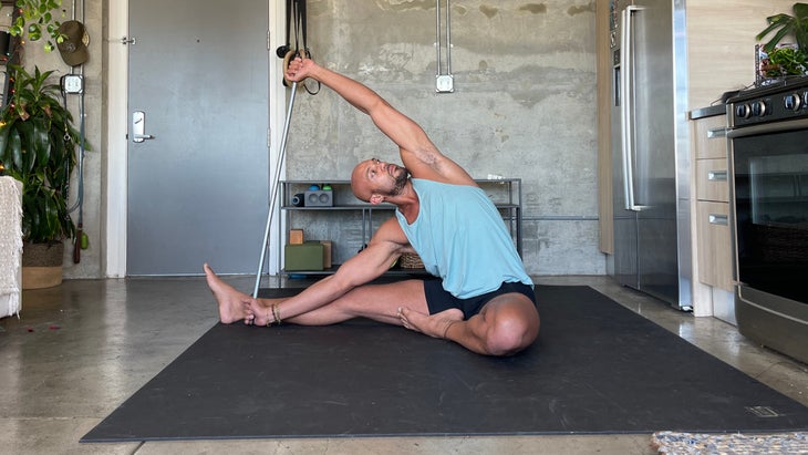Parivrtta Janu Sirsasana. Yoga teacher Rocky Heron wears a light blue tank and dark shorts. He is standing on a dark/black mat with a concrete wall in the background.