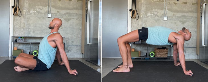 Reverse table. Yoga teacher Rocky Heron wears a light blue tank and dark shorts. He is standing on a dark/black mat with a concrete wall in the background.