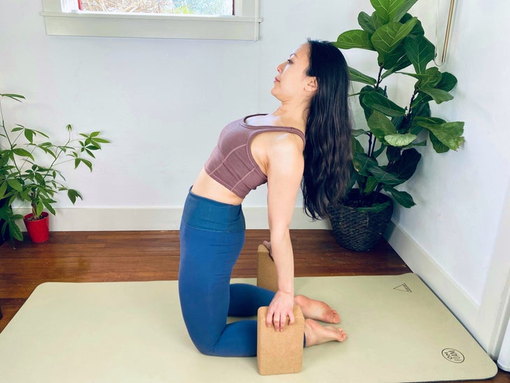 An Asian woman practices Camel Pose. She wears a brown top and blue tights, and is kneeling on a light green yoga mat. She is in a white room with large plants in the background.