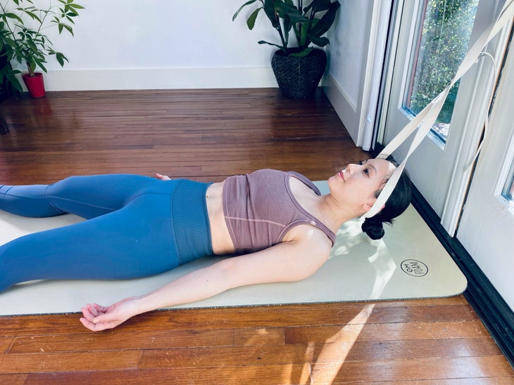 An Asian woman uses a yoga strap attached to a doorknob to create neck traction. She wears a brown top and blue tights, and is lying on a light green yoga mat with her head held by the stap. She is in a white room with large plants in the background.