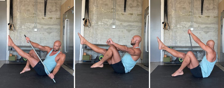 Rocky Heron demonstrates a variation of Navasana using a dowel. He is wearing a light blue tank and dark shorts, sitting on a black mat against a concrete background.