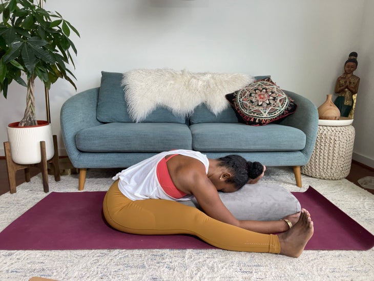 Faith Hunter practices supported Forward Fold with a bolster. She is wearing a white shirt and gold tights and sits on a burgundy mat. In the background is a blue-gray sofa with an embroidered pillow and a large green plant in a white pot.