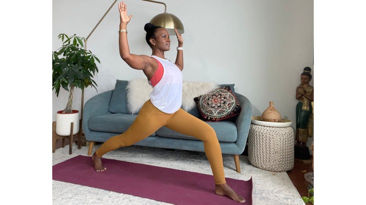 Faith Hunter practices a High Lunge with Cactus Arms. She is wearing a white shirt and gold tights and sits on a burgundy mat. In the background is a blue-gray sofa with an embroidered pillow and a large green plant in a white pot.