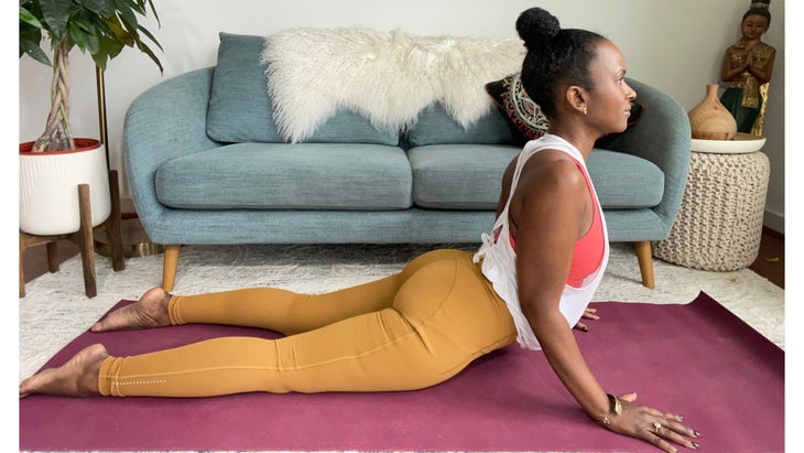 Faith Hunter practices King Cobra pose. She is wearing a white shirt and gold tights and sits on a burgundy mat. In the background is a blue-gray sofa with an embroidered pillow and a large green plant in a white pot.