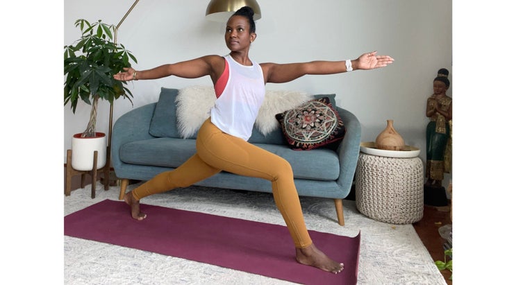 Faith Hunter practices a High Lunge with a Twist. She is wearing a white shirt and gold tights and sits on a burgundy mat. In the background is a blue-gray sofa with an embroidered pillow and a large green plant in a white pot.