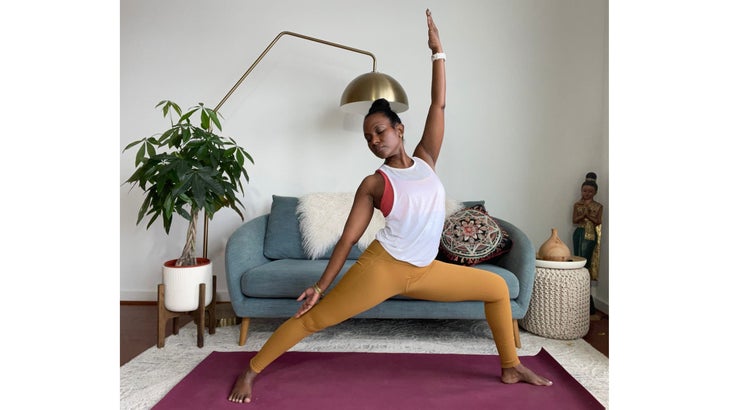 Faith Hunter practices Peaceful Warrior Pose. She is wearing a white shirt and gold tights and sits on a burgundy mat. In the background is a blue-gray sofa with an embroidered pillow and a large green plant in a white pot.