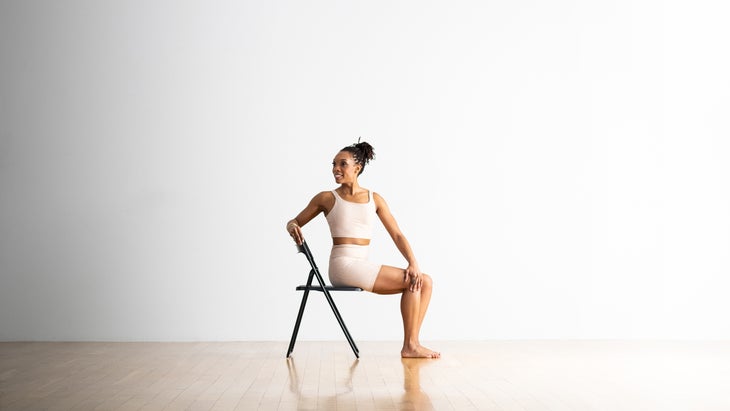 Bharadvajasana, Bharadvaja's twist on a chair. She wears white yoga shorts and top and sits against a white background