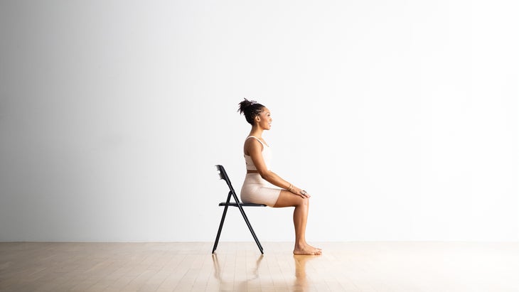 Easy Pose, Sukhasana, on a chair. She wears white yoga shorts and top and sits against a white background